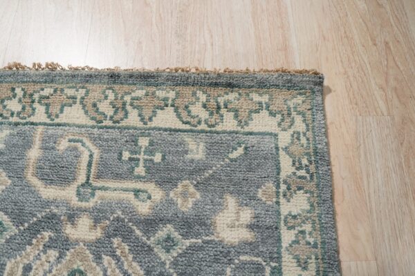 Textured blue-gray rug featuring a traditional border design in cream and teal on light wood flooring.