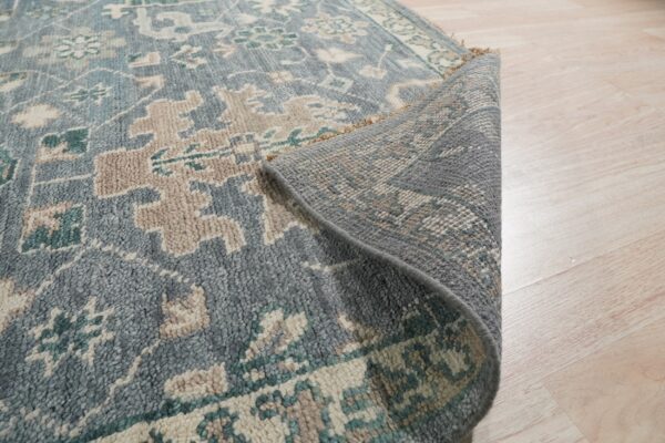 Dusty blue rug with cream, taupe, and teal traditional patterns curled up on pale wooden flooring.
