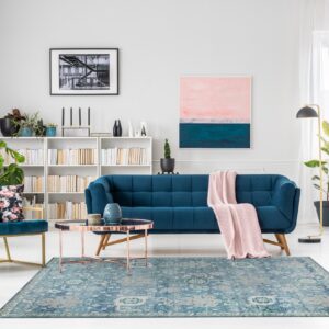 Muted blue traditional rug centers a room with a teal sofa, pink throw, and white wood flooring.