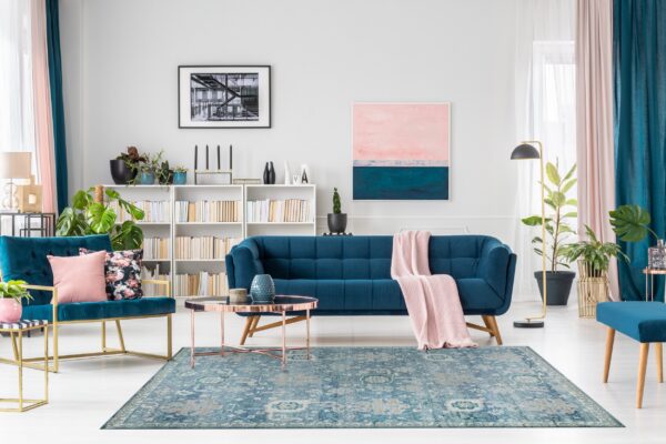 Muted blue traditional rug centers a room with a teal sofa, pink throw, and white wood flooring.