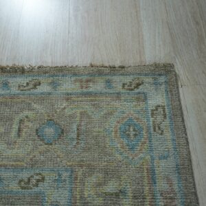 Textured rug corner displaying taupe, blue, yellow, and pink geometric patterns on light blonde wood planks.