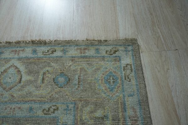 Textured rug corner displaying taupe, blue, yellow, and pink geometric patterns on light blonde wood planks.