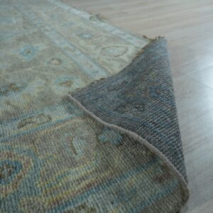 Muted gray and blue patterned rug with a medium pile, curled back to show the woven underside on wood flooring.