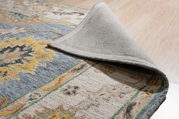 A high-pile rug with blue, cream, and yellow traditional patterns is curled back, showing its gray backing on light wood floor.