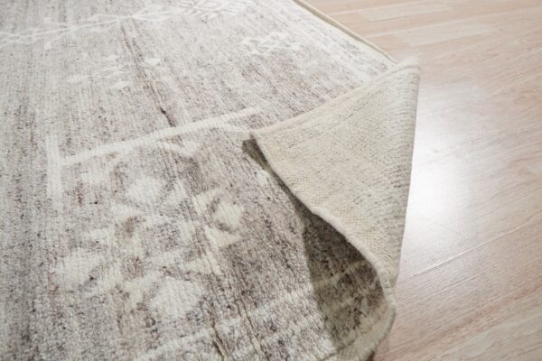 A textured off-white rug with brown speckling is peeled back on light wood plank flooring.