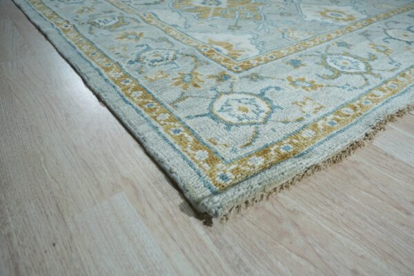 Textured rug corner in light blue, gold, and white with a traditional motif on pale wood flooring.