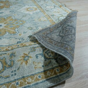 Blue, cream, and gold patterned rug surface and gray backing shown on light wood planks.