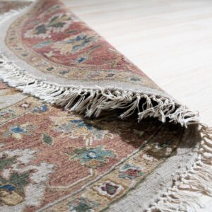 Muted terracotta, beige, and green floral rug with long tassels is partially curled on bright blonde wood floors.