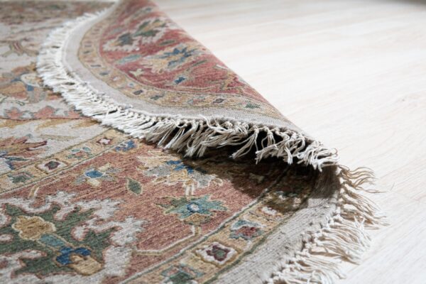 Muted terracotta, beige, and green floral rug with long tassels is partially curled on bright blonde wood floors.