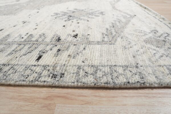 Textured, cream-colored rug with gray abstract motifs and speckling rests on light wooden flooring.