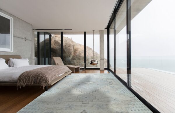 A low-pile gray and light blue geometric rug grounds a modern bedroom with concrete walls and window views.