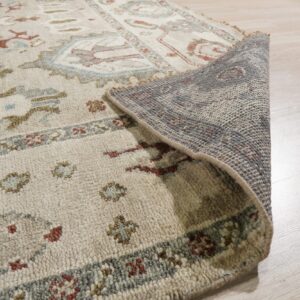 Close-up of a textured beige rug with traditional patterns, showing the gray backing on light wood flooring.
