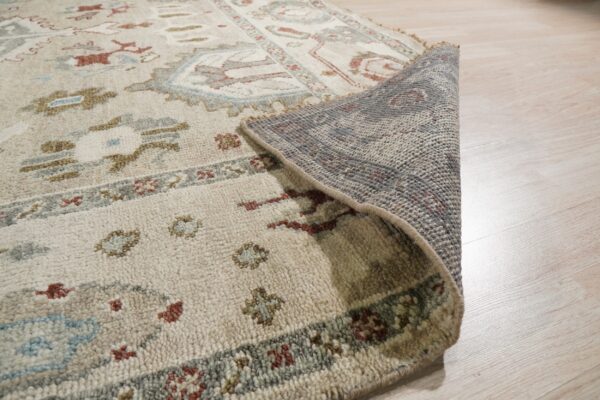 Close-up of a textured beige rug with traditional patterns, showing the gray backing on light wood flooring.