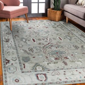 Pale gray patterned rug with maroon and teal designs on wood floor between a pink chair and gray sofa.
