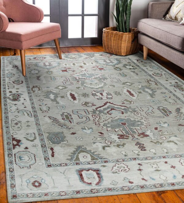 Pale gray patterned rug with maroon and teal designs on wood floor between a pink chair and gray sofa.