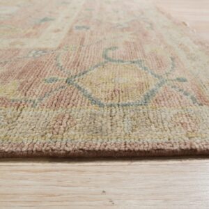 Angled view of a medium pile rug featuring rosy pink, cream, and blue designs on blonde wood flooring.