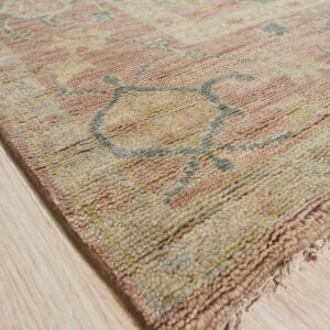Chunky pile rug in rose, beige, and blue-gray geometric patterns resting on light wood flooring.