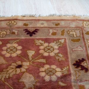 Close-up of a rose and tan traditional floral rug corner with fringe on light plank flooring.