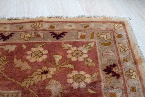Close-up of a rose and tan traditional floral rug corner with fringe on light plank flooring.