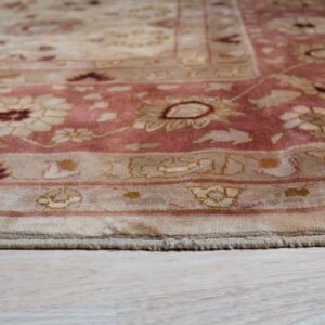 Low-angle view of a low-pile traditional rug border in rose and beige on light wood planks.