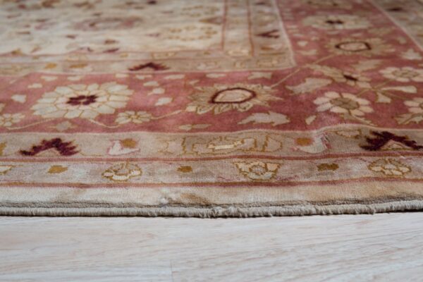 Low-angle view of a low-pile traditional rug border in rose and beige on light wood planks.