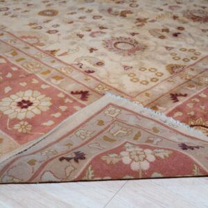 Traditional floral rug in beige and pink colors, with a corner folded over light wood floor.