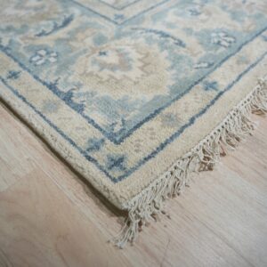 Close up of a cream and blue traditional rug with fringe on light wood flooring.