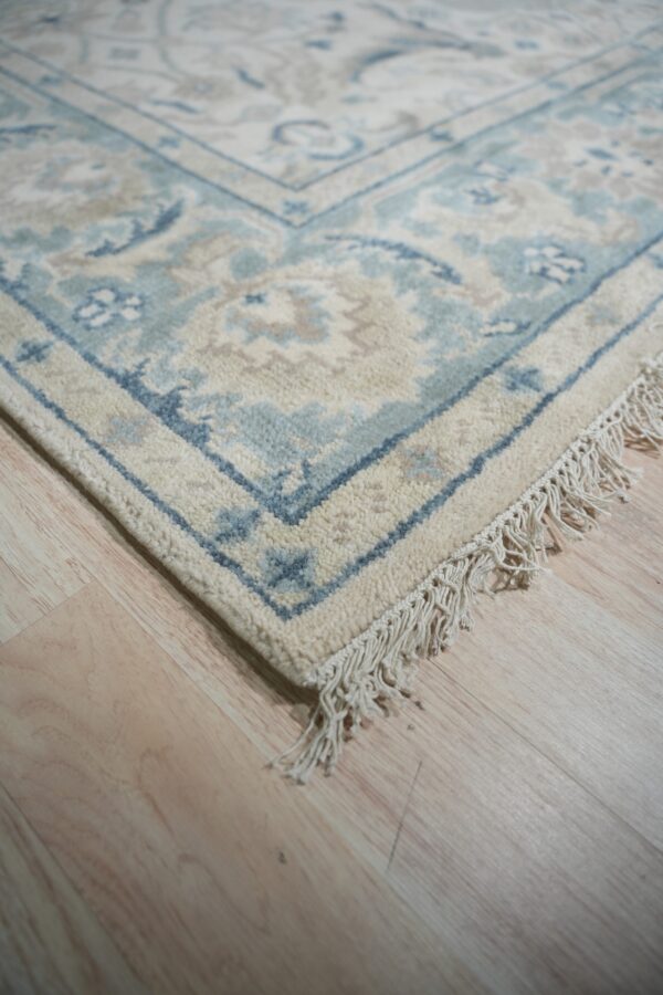 Close up of a cream and blue traditional rug with fringe on light wood flooring.
