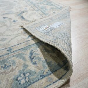 Close-up of a cream and blue patterned rug with low pile, partially rolled up on light wood flooring.