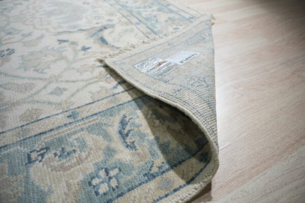 Close-up of a cream and blue patterned rug with low pile, partially rolled up on light wood flooring.