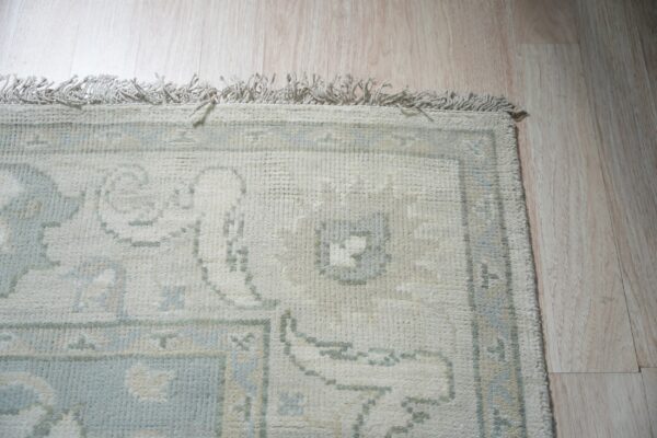 Muted cream and light blue traditional pattern rug with fringes on light blonde plank flooring.