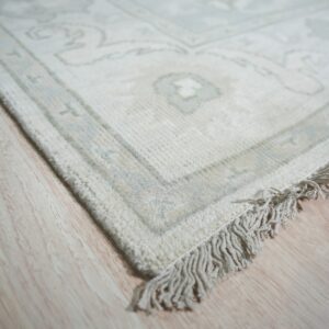 Low-contrast ivory rug showing ornate pattern and fringes on pale blonde plank flooring.