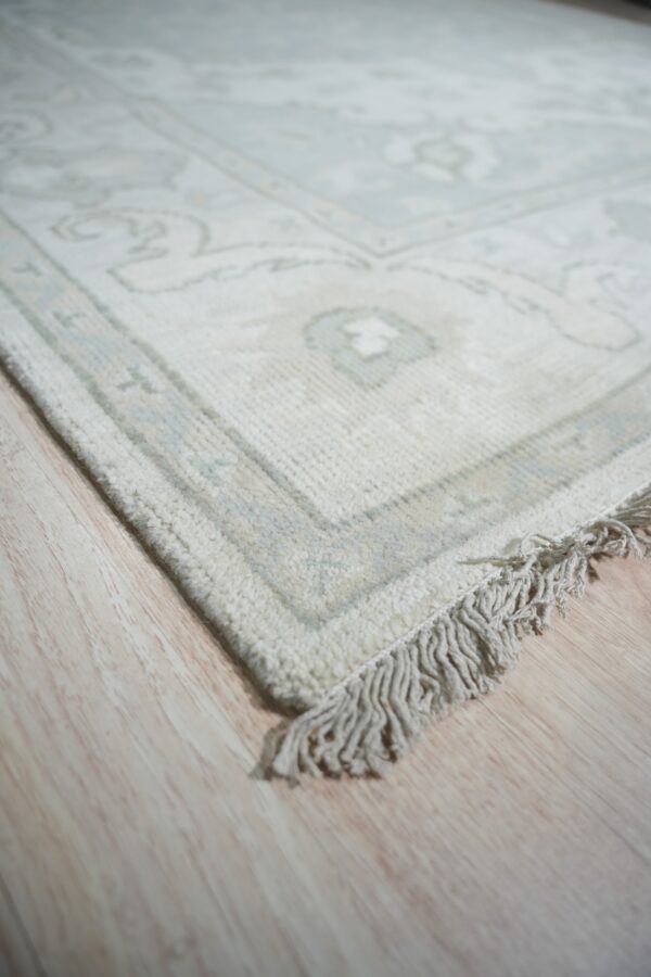 Low-contrast ivory rug showing ornate pattern and fringes on pale blonde plank flooring.