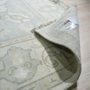 Pale blue and cream patterned rug corner curled back, showing the backing, fringe, and label on wood flooring.