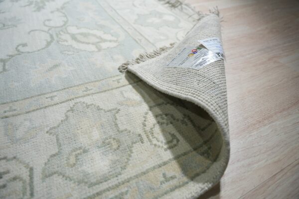 Pale blue and cream patterned rug corner curled back, showing the backing, fringe, and label on wood flooring.