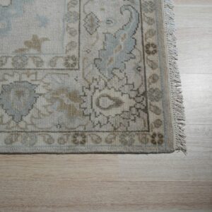 The border of a textured rug in cream, light blue, and brown lies on light blonde wood flooring.