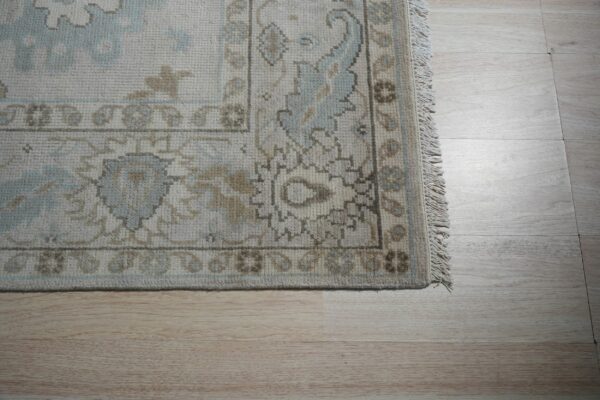 The border of a textured rug in cream, light blue, and brown lies on light blonde wood flooring.