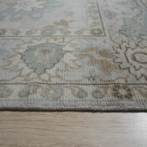 Muted pale blue and tan low pile rug with traditional designs rests on light wood flooring.