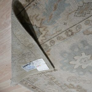 Close-up of a rolled low-pile beige rug with blue and brown traditional patterns on light wood flooring.