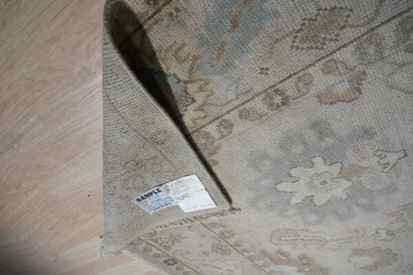 Close-up of a rolled low-pile beige rug with blue and brown traditional patterns on light wood flooring.