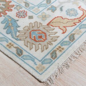 Corner of a cream rug featuring traditional blue, tan, and terracotta floral motifs on light wood flooring.