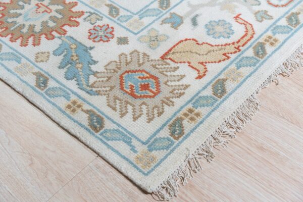 Corner of a cream rug featuring traditional blue, tan, and terracotta floral motifs on light wood flooring.