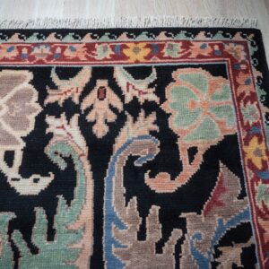 Black rug with beige, red, and sage floral patterns and fringe lies on light wood planks.