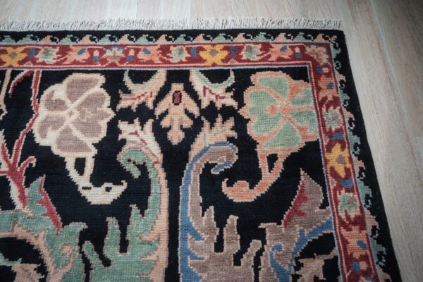 Black rug with beige, red, and sage floral patterns and fringe lies on light wood planks.