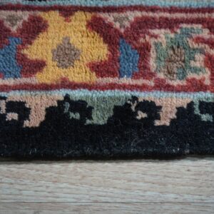 Close-up of a medium pile rug border in red, gold, blue, and black on light wood flooring.
