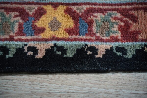 Close-up of a medium pile rug border in red, gold, blue, and black on light wood flooring.
