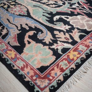 A black, plush rug with multicolored traditional botanical designs and fringe rests on light wood flooring.