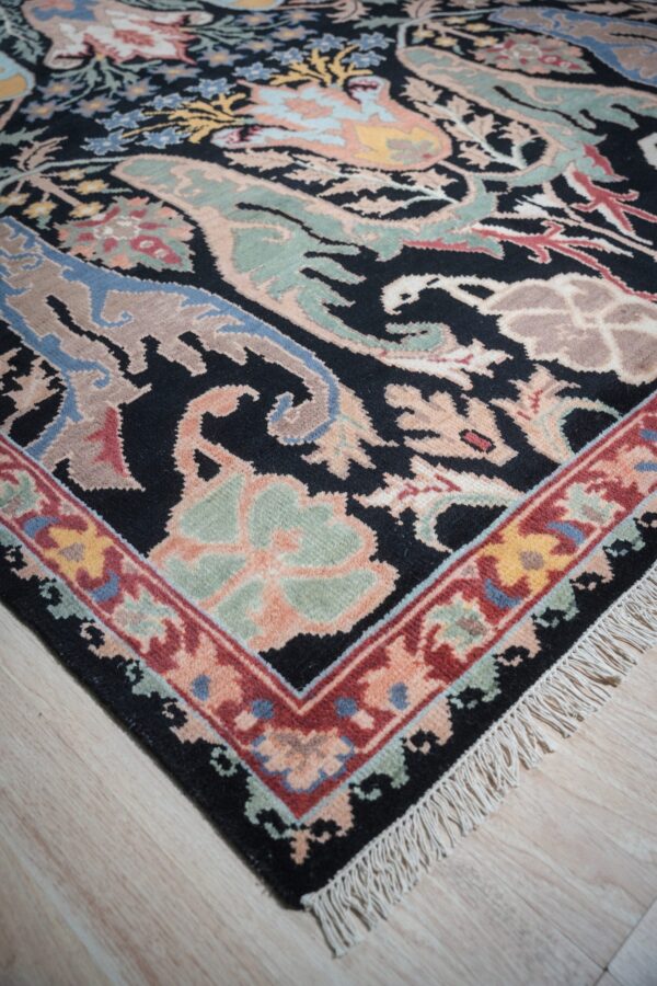 A black, plush rug with multicolored traditional botanical designs and fringe rests on light wood flooring.