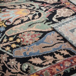 A black rug with multicolored floral patterns, including blue and sage, is rolled up on light wood floor.