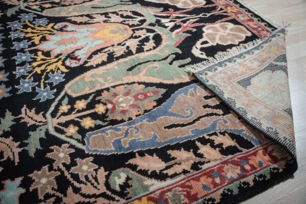 A black rug with multicolored floral patterns, including blue and sage, is rolled up on light wood floor.